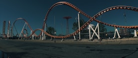 A large roller coaster with orange tracks and white supports stretches across an amusement park setting. The tracks loop and twist in intricate patterns against a cloudy sky. In the background, there are other amusement rides and structures, as well as trees lining the area.