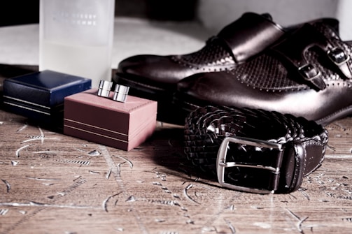 A pair of polished dark brown formal shoes is placed on a wooden surface. Next to them are two small, closed boxes, one blue and one red. There's also a braided dark brown belt with a silver buckle. In the background, there is a partially visible frosted glass bottle.