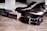 A pair of polished dark brown formal shoes is placed on a wooden surface. Next to them are two small, closed boxes, one blue and one red. There's also a braided dark brown belt with a silver buckle. In the background, there is a partially visible frosted glass bottle.