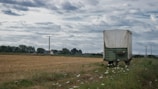 A rural landscape with a large, weathered green and white trailer parked on the edge of a harvested field. The field is surrounded by power lines and scattered houses in the distance under a cloudy sky. Wildflowers grow alongside the path next to the trailer.