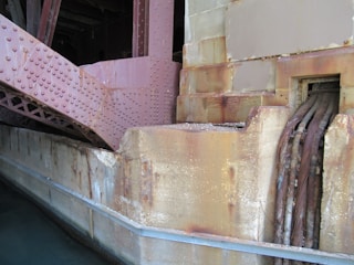 A close-up view of a large industrial structure made of metal and concrete. The metal beams are rust-colored with numerous rivets and connect to a stone or concrete wall with stained streaks. Thick cables are exposed, emerging from a small opening in the wall.