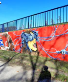 A vibrant graffiti mural on a concrete wall depicting a woman's face wearing a virtual reality headset connected with multiple wires. Her expression is intense and dramatic, set against a bright red background. The image takes place outdoors with grass and a blue sky visible, and a blue railing above the wall.