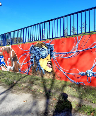 A vibrant graffiti mural on a concrete wall depicting a woman's face wearing a virtual reality headset connected with multiple wires. Her expression is intense and dramatic, set against a bright red background. The image takes place outdoors with grass and a blue sky visible, and a blue railing above the wall.