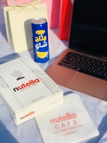 A white box with 'Nutella' branding sits on a light surface, accompanied by a can of cold beverage with Arabic text and a lemon graphic. Beside the box lies a napkin with 'Nutella Cafe' branding, and a laptop with a rose gold casing partially visible. In the background, there are two paper bags, one white, the other red, slightly out of focus.