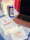 A white box with 'Nutella' branding sits on a light surface, accompanied by a can of cold beverage with Arabic text and a lemon graphic. Beside the box lies a napkin with 'Nutella Cafe' branding, and a laptop with a rose gold casing partially visible. In the background, there are two paper bags, one white, the other red, slightly out of focus.