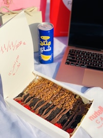 A dessert box containing layered cake slices topped with chocolate, cookie crumbs, and colorful crumbles is placed on a table. Beside it, there's a blue and yellow can of drink with Arabic text. A rose gold laptop is partially visible, and a napkin with 'Nutella' branding is on the side. In the background, there are gift bags and a card with dates written in red.