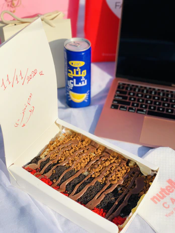 A dessert box containing layered cake slices topped with chocolate, cookie crumbs, and colorful crumbles is placed on a table. Beside it, there's a blue and yellow can of drink with Arabic text. A rose gold laptop is partially visible, and a napkin with 'Nutella' branding is on the side. In the background, there are gift bags and a card with dates written in red.