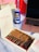 A dessert box containing layered cake slices topped with chocolate, cookie crumbs, and colorful crumbles is placed on a table. Beside it, there's a blue and yellow can of drink with Arabic text. A rose gold laptop is partially visible, and a napkin with 'Nutella' branding is on the side. In the background, there are gift bags and a card with dates written in red.