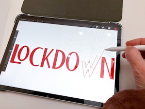 A tablet displays the word 'LOCKDOWN' in bold red letters. A hand holding a stylus is interacting with the screen, aligning or drawing the final letter 'N'. The background is mostly white, and the tablet is placed on a light-colored surface.