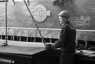 A worker wearing protective gear, including a helmet and gloves, operates or supervises a large industrial machine. The setting has a heavy, metallic texture with chains and machinery parts visible. The environment appears to be a factory or industrial workspace, evidenced by the presence of metal and technical equipment.