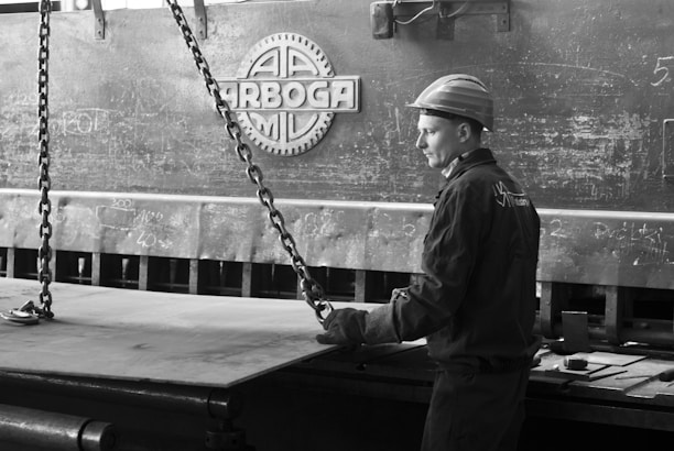 A worker wearing protective gear, including a helmet and gloves, operates or supervises a large industrial machine. The setting has a heavy, metallic texture with chains and machinery parts visible. The environment appears to be a factory or industrial workspace, evidenced by the presence of metal and technical equipment.