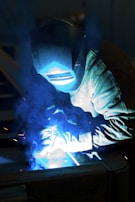 A welder is working, with bright blue sparks and smoke illuminating the scene. The person is wearing protective gear, including a welding helmet and gloves, focusing on metalwork.