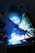 A welder is working, with bright blue sparks and smoke illuminating the scene. The person is wearing protective gear, including a welding helmet and gloves, focusing on metalwork.
