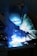A welder is working, with bright blue sparks and smoke illuminating the scene. The person is wearing protective gear, including a welding helmet and gloves, focusing on metalwork.