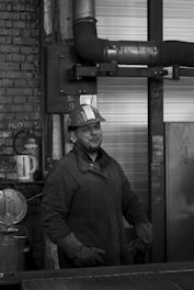 A worker wearing a hard hat stands in an industrial setting, surrounded by machinery and metal pipes. The image is in black and white, giving it a timeless, gritty feel. The worker appears confident and experienced, with a slight smile on his face.