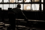 A silhouette of a man wearing a hard hat and working inside a dimly lit industrial space. He is facing a large, dirty window with several broken panes and is handling a heavy object on a table or machinery. Chains hang from above, adding to the industrial atmosphere.