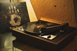 A vintage turntable is playing a record in a dimly lit room. It features a wooden base and a sleek tonearm. Next to the turntable, there's a vinyl album cover propped up with a handprint design and the text 'Beat Time.' The walls have a textured appearance, contributing to a nostalgic ambiance.