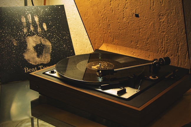 A vintage turntable is playing a record in a dimly lit room. It features a wooden base and a sleek tonearm. Next to the turntable, there's a vinyl album cover propped up with a handprint design and the text 'Beat Time.' The walls have a textured appearance, contributing to a nostalgic ambiance.