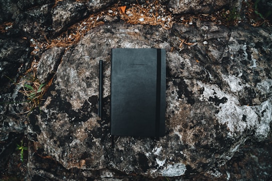 A black notebook and pen are placed on a textured rock surface. The rock has various shades of gray and brown, with small patches of green moss and pine needles scattered around. The scene suggests a natural, outdoor setting.