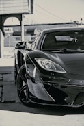 A sleek, high-end sports car is parked in an industrial area with a chain-link fence and a FedEx logo visible in the background. The image focuses on the front side of the car, prominently displaying its glossy finish and modern aerodynamic design.