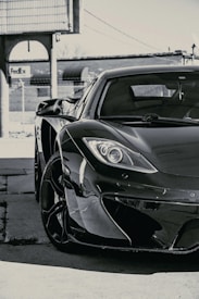 A sleek, high-end sports car is parked in an industrial area with a chain-link fence and a FedEx logo visible in the background. The image focuses on the front side of the car, prominently displaying its glossy finish and modern aerodynamic design.