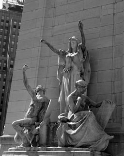 A detailed stone sculpture featuring three figures in classical poses, set against an engraved architectural backdrop. The central figure appears to be a robed woman with arms raised, flanked by two seated figures. The one on the left looks upwards, while the right figure is holding a tablet.