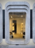 A storefront window framed with layered, rounded marble slabs reveals an interior space featuring abstract black and white artworks. The lighting inside highlights the art pieces and enhances the modern aesthetic. The display is minimalistic with clean lines and a neutral color palette.