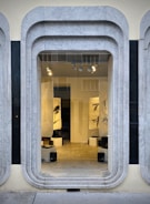 A storefront window framed with layered, rounded marble slabs reveals an interior space featuring abstract black and white artworks. The lighting inside highlights the art pieces and enhances the modern aesthetic. The display is minimalistic with clean lines and a neutral color palette.