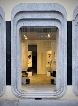 A storefront window framed with layered, rounded marble slabs reveals an interior space featuring abstract black and white artworks. The lighting inside highlights the art pieces and enhances the modern aesthetic. The display is minimalistic with clean lines and a neutral color palette.