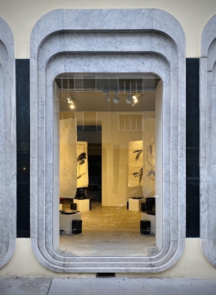 A storefront window framed with layered, rounded marble slabs reveals an interior space featuring abstract black and white artworks. The lighting inside highlights the art pieces and enhances the modern aesthetic. The display is minimalistic with clean lines and a neutral color palette.