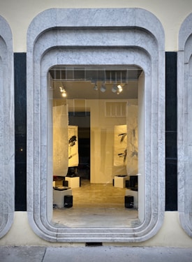 A storefront window framed with layered, rounded marble slabs reveals an interior space featuring abstract black and white artworks. The lighting inside highlights the art pieces and enhances the modern aesthetic. The display is minimalistic with clean lines and a neutral color palette.