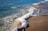 A person holding a surfboard is about to enter the ocean from a sandy beach. The waves gently crash onto the shoreline, creating foam where the water meets the sand. The individual is wearing a wetsuit, and the scene suggests a sunny day by the sea.