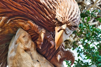 A detailed wooden sculpture of an eagle with a fierce expression, showcasing intricate carving on the feathers. The claws are prominently depicted, adding to the lifelike appearance. The background includes some green leaves.