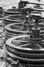 A series of industrial metal pulleys and cables in a black-and-white setting, showcasing a focus on machinery components. The metal appears worn and weathered, suggesting usage over time.