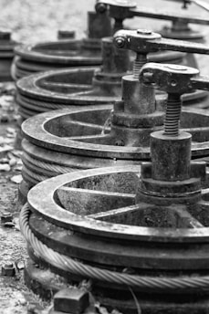 A series of industrial metal pulleys and cables in a black-and-white setting, showcasing a focus on machinery components. The metal appears worn and weathered, suggesting usage over time.