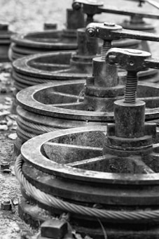 A series of industrial metal pulleys and cables in a black-and-white setting, showcasing a focus on machinery components. The metal appears worn and weathered, suggesting usage over time.