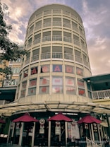 A tall, cylindrical building with multiple stories featuring a series of horizontal, white railings. The lower half displays windows with red text advertising body therapy, massage, and bone alignment. Below the windows, a row of maroon umbrellas shades an outdoor seating area alongside a restaurant entrance.