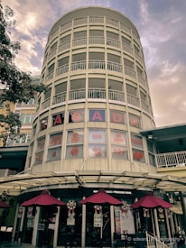 A tall, cylindrical building with multiple stories featuring a series of horizontal, white railings. The lower half displays windows with red text advertising body therapy, massage, and bone alignment. Below the windows, a row of maroon umbrellas shades an outdoor seating area alongside a restaurant entrance.