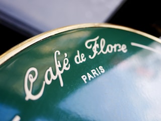 A close-up view of a green circular sign featuring the words 'Café de Flore' and 'Paris' in white, elegant lettering.