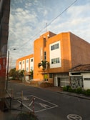 A large, brick building with modern architectural elements is situated on a quiet street. The facade features large windows and a few palm trees line the entrance. The road in front has designated parking spaces and some caution signs, with a clear sky overhead.