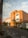 A large, brick building with modern architectural elements is situated on a quiet street. The facade features large windows and a few palm trees line the entrance. The road in front has designated parking spaces and some caution signs, with a clear sky overhead.