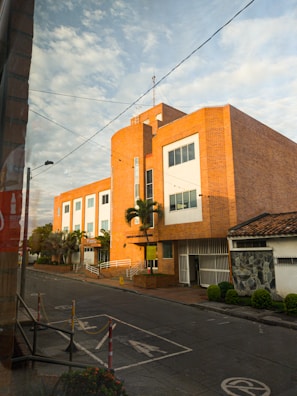 A large, brick building with modern architectural elements is situated on a quiet street. The facade features large windows and a few palm trees line the entrance. The road in front has designated parking spaces and some caution signs, with a clear sky overhead.