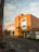 A large, brick building with modern architectural elements is situated on a quiet street. The facade features large windows and a few palm trees line the entrance. The road in front has designated parking spaces and some caution signs, with a clear sky overhead.