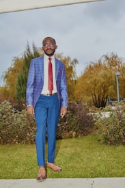 A man dressed in a stylish blue blazer with a check pattern, dark blue pants, a white shirt, and a red tie. He stands outdoors on a grassy area with trimmed bushes and trees displaying autumnal colors in the background. The sky is overcast, adding a neutral backdrop.