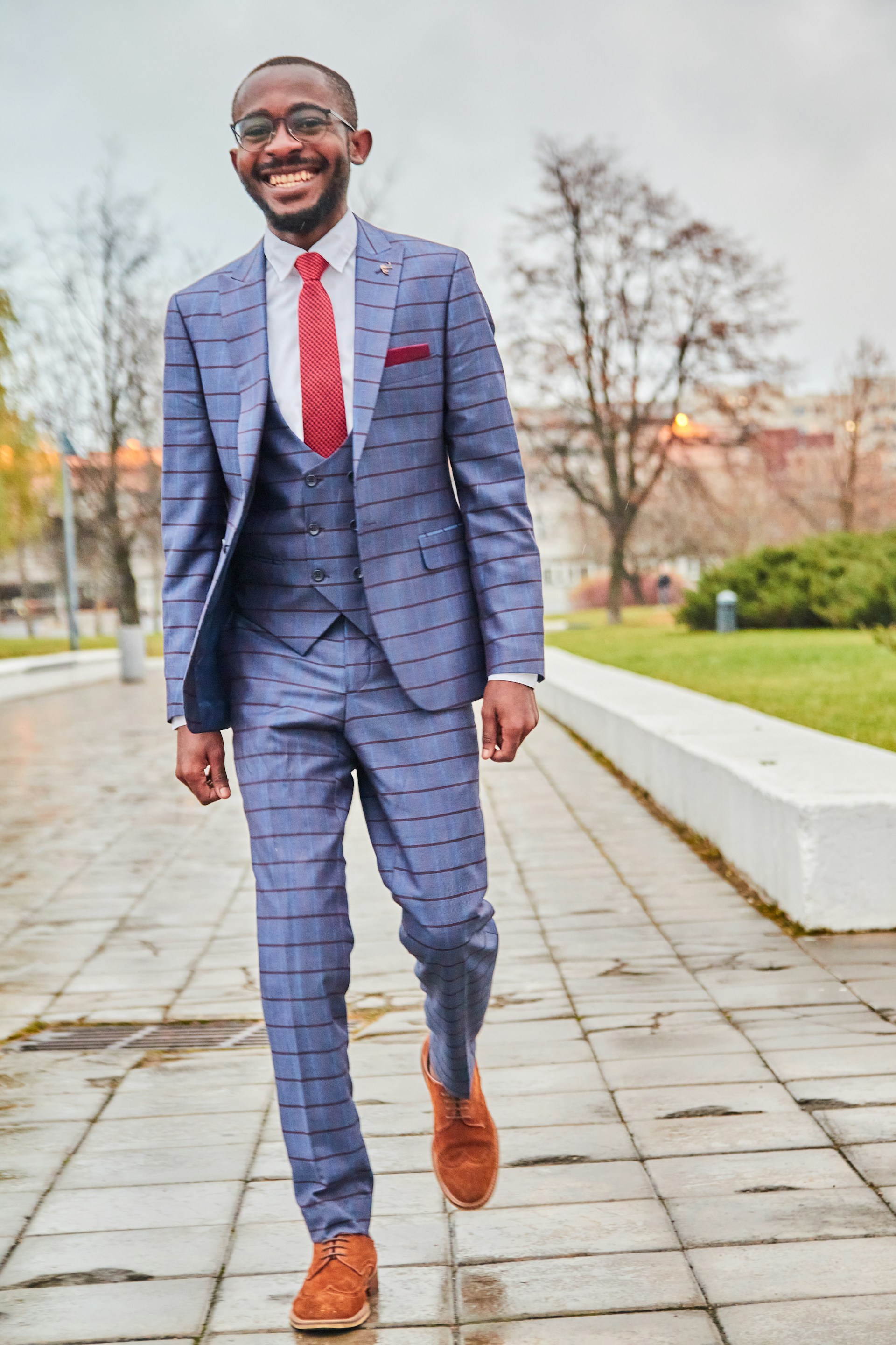 A person wearing a stylish, fitted blue suit with red pinstripes and a red tie, smiling broadly while walking down a paved path in an outdoor setting. The surroundings are park-like with some trees, a grass area, and a cloudy sky.