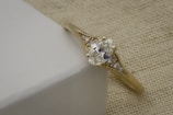 A gold ring with a central, large, round diamond flanked by smaller diamonds is resting on a light-colored textured surface. The setting is intricate, showcasing fine details and craftsmanship.