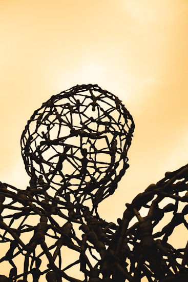 A metal sculpture composed of interconnected humanoid figures forms a lattice structure that resembles a head and shoulders against a warm, yellowish background.