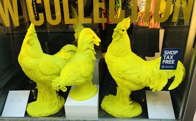 Three large, bright yellow rooster sculptures are displayed in a shop window. The background features text on the glass and colorful clothing. Two price tags are visible on the white surfaces near the sculptures.