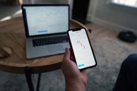 A person holds a smartphone displaying a stock market trading app with candlestick charts. A laptop is on a wooden table with a similar financial chart, indicating active trading or analysis. The setting suggests a home office or casual workspace.
