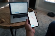 A person holds a smartphone displaying a stock market trading app with candlestick charts. A laptop is on a wooden table with a similar financial chart, indicating active trading or analysis. The setting suggests a home office or casual workspace.